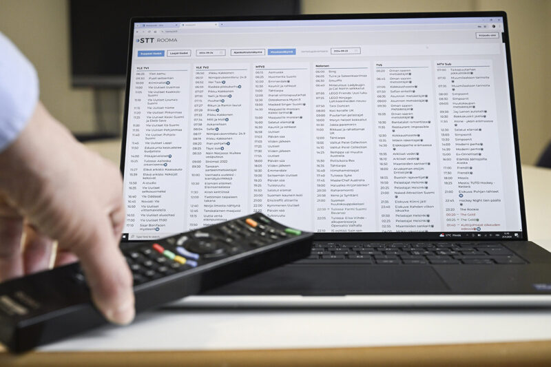 STT Rooma -ohjelmatietodatapalvelun käyttöliittymä tietokoneen näytöllä, etualalla naisen kädessä kaukosäädin.