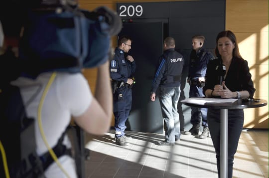 Toimittaja valmistautuu puhumaan tv-kameralle Helsingin käräjäoikeudessa, taustalla poliiseja salin ovella.