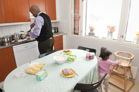 Tummaihoinen isä laittaa ruokaa keittiössä pienen tyttären katsellessa.