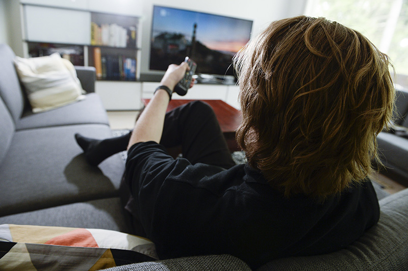 Mies käyttää kaukosäädintä katsoessaan televisiota kotisohvalla.